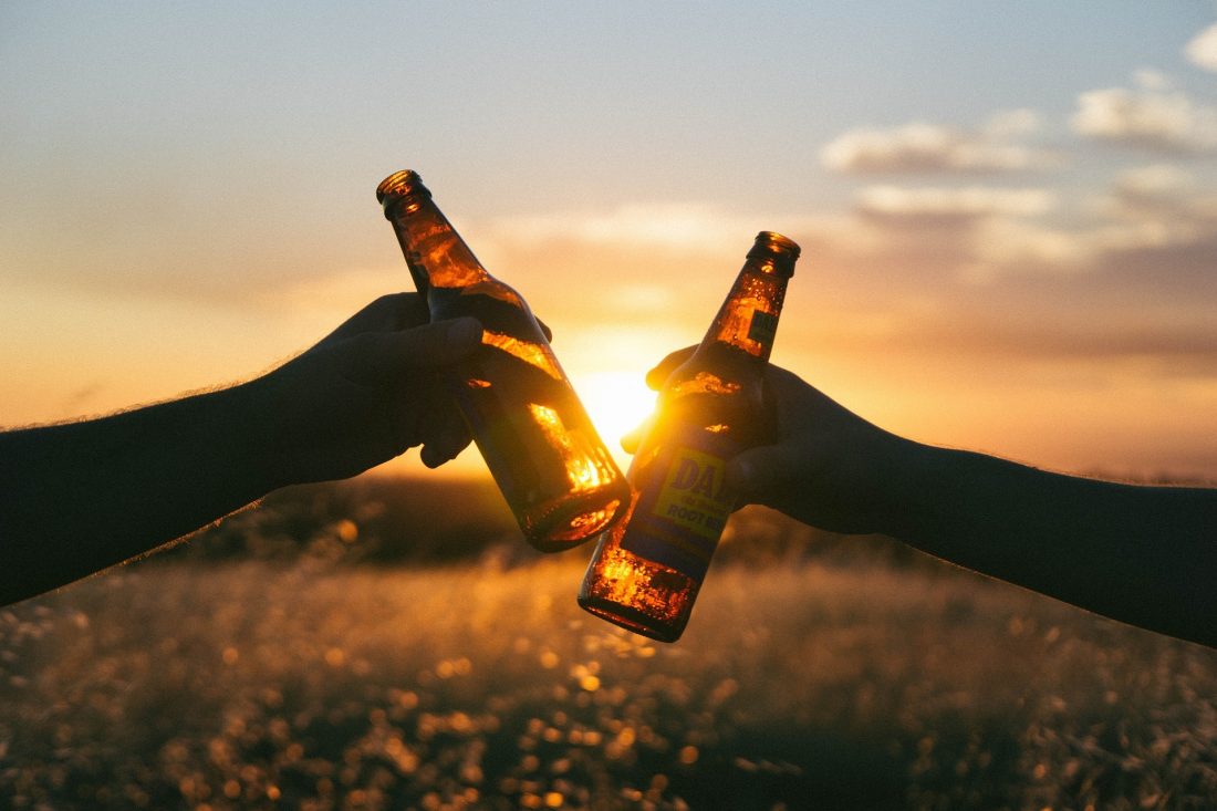 Foto von zwei Händen, die bei Sonnenuntergang mit zwei Flaschen Bier anstoßen