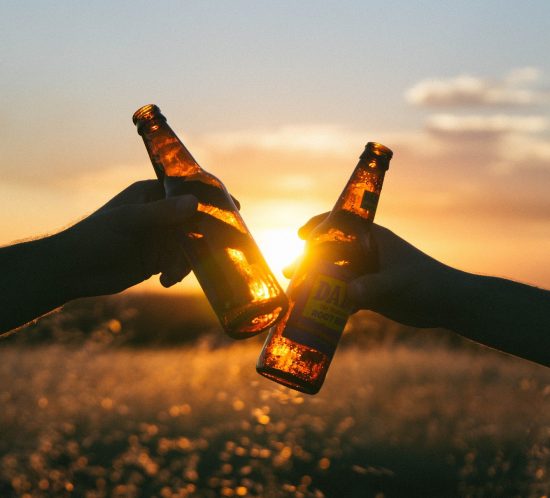 Foto von zwei Händen, die bei Sonnenuntergang mit zwei Flaschen Bier anstoßen